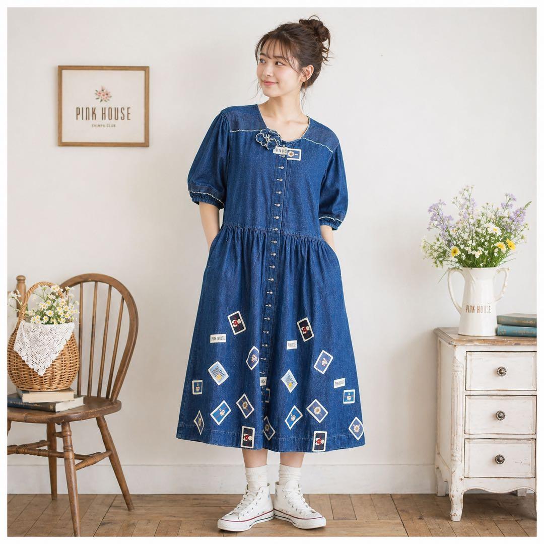 Model wearing the long blue denim PINK HOUSE dress with front buttons and multiple decorative patches, photographed in a room.
