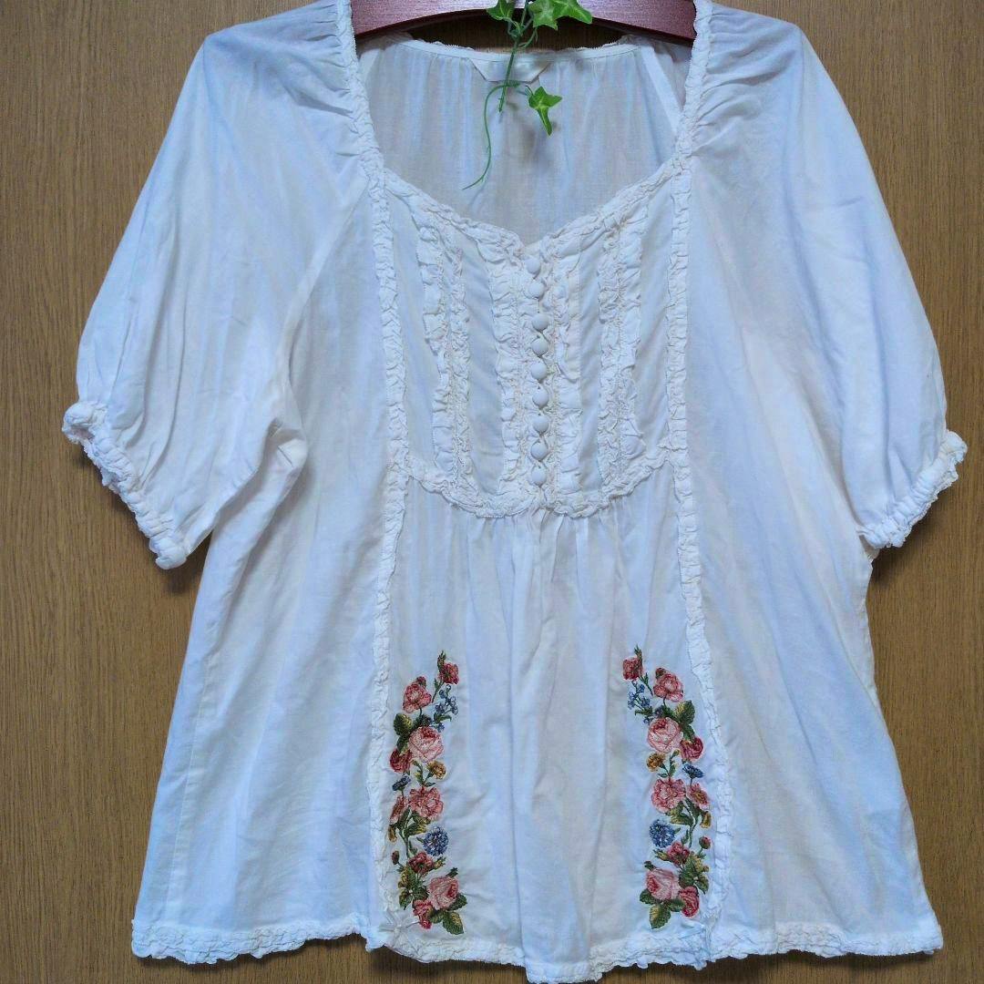 White A-line blouse with rose embroidery hanging on a wooden hanger against a wooden background.