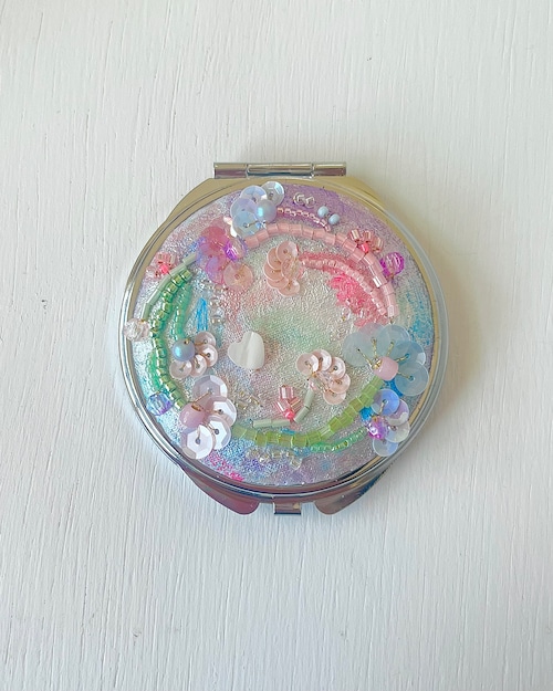 A round compact mirror with a decorative pastel sequin-and-bead cover placed on a white surface.