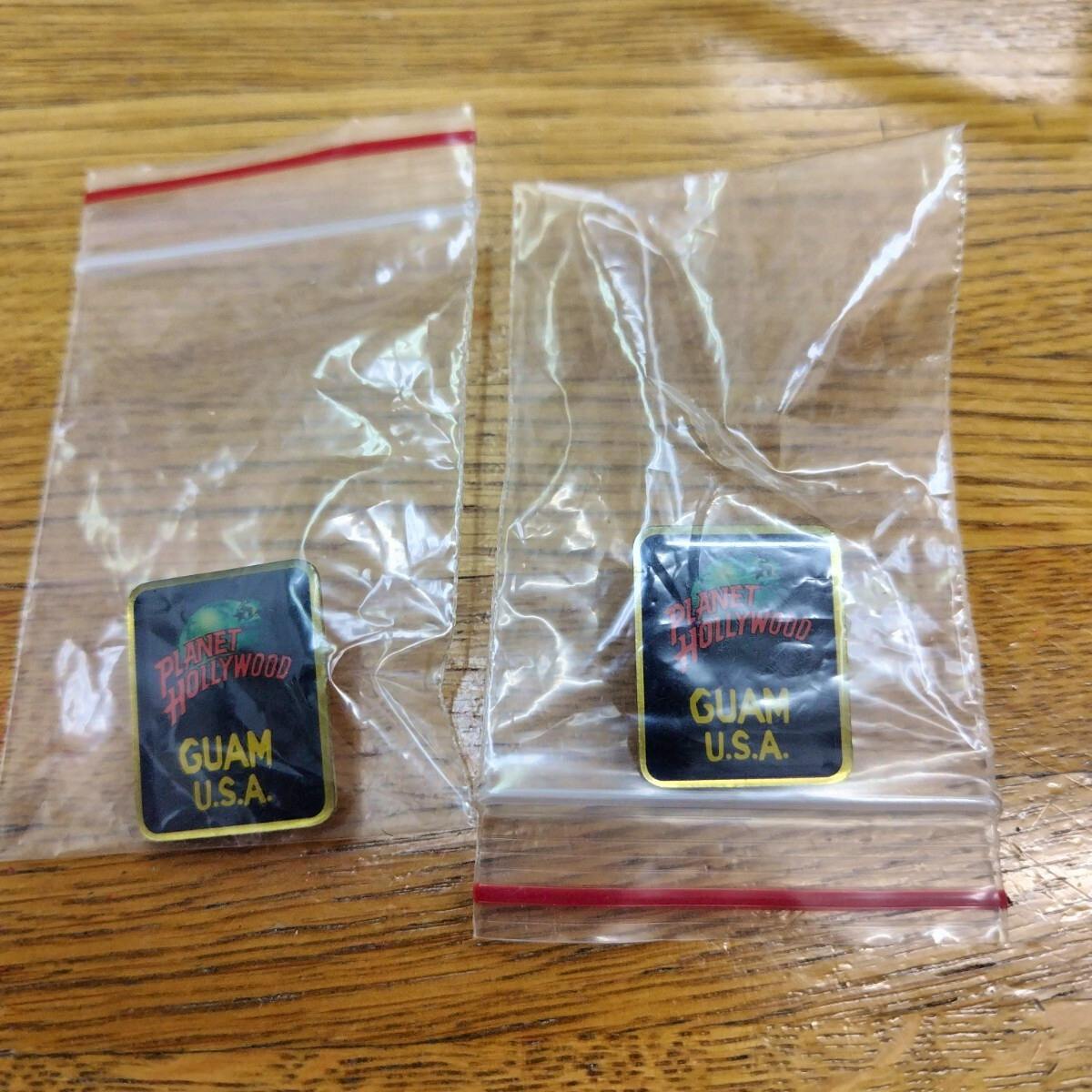 Two Planet Hollywood Guam U.S.A. pin badges in small plastic bags placed on a wooden surface.