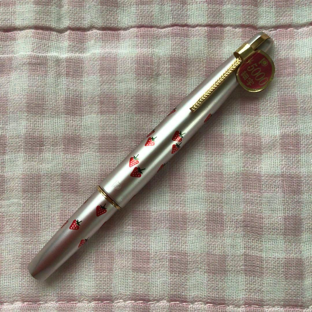 Strawberry-patterned capped fountain pen with a gold clip and round tag, on pink checked cloth.