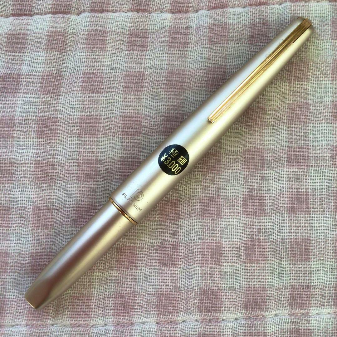 Closed Platinum short fountain pen with a metallic cap and clip, showing a round sticker reading "極細 ¥3,000", lying on a pink checkered cloth.