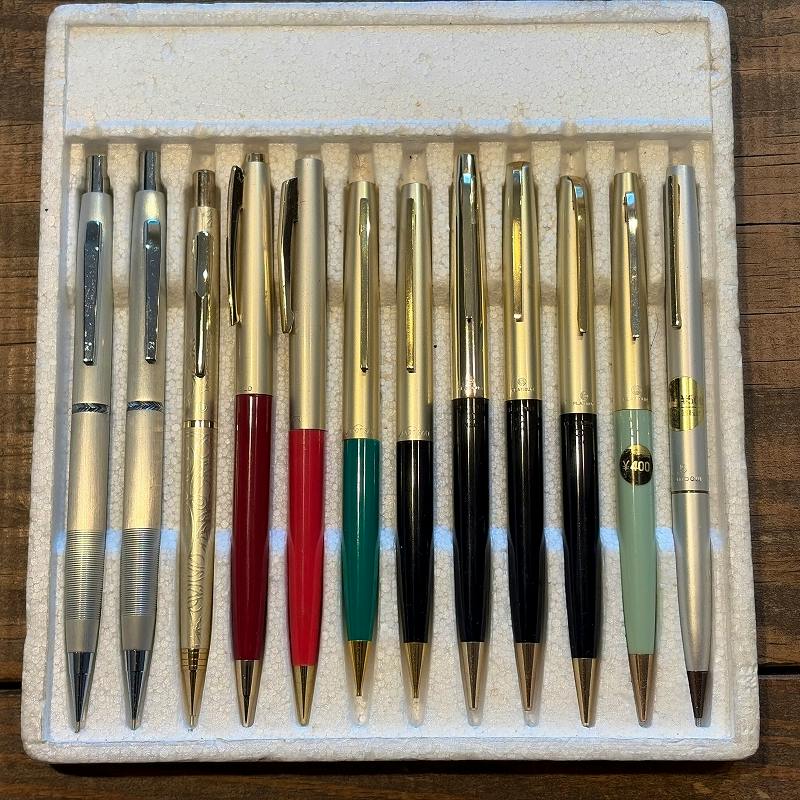 Top-down photo of twelve mechanical pencils and similar click/rotary pencils arranged side-by-side in a foam tray on a wooden surface.