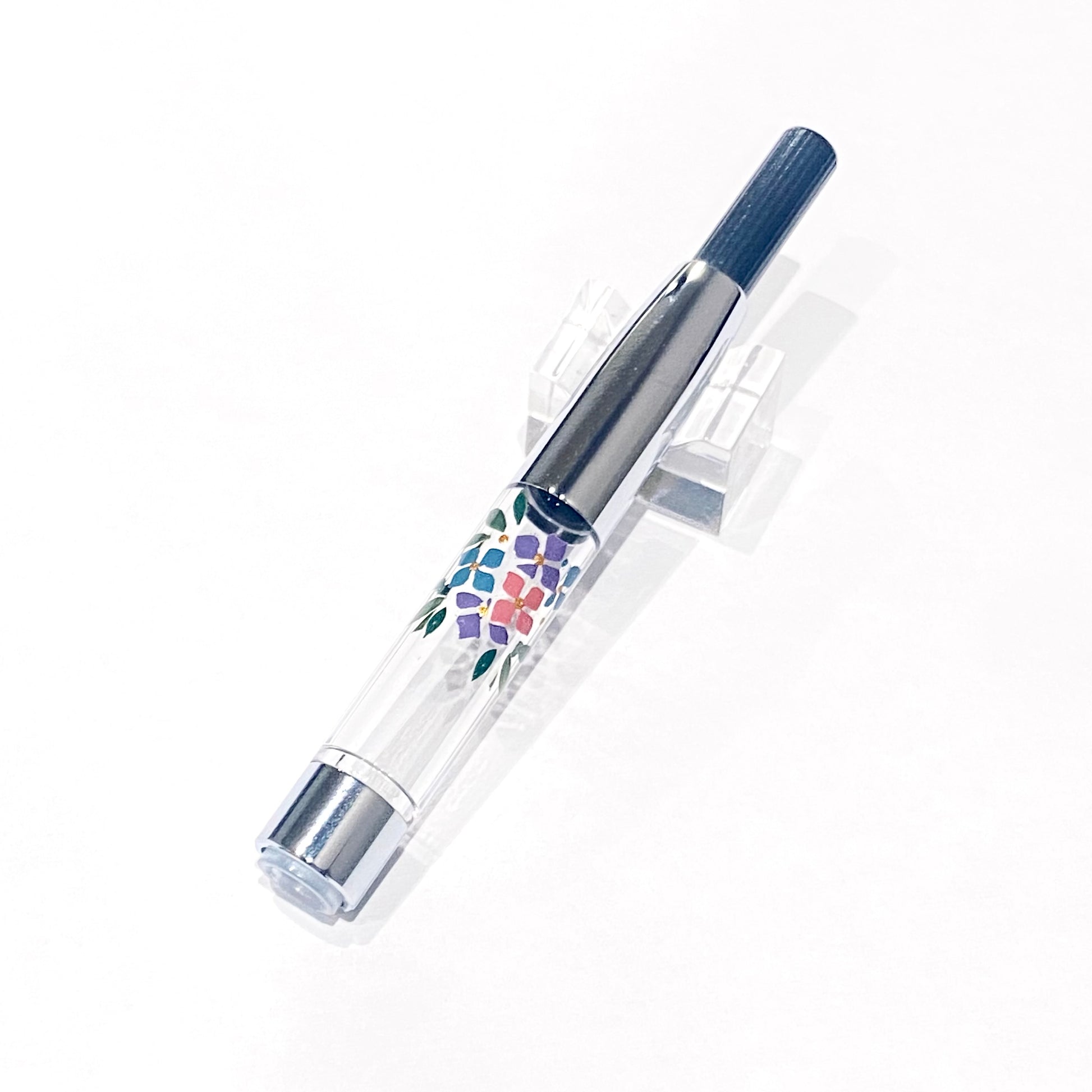 Ink converter for a fountain pen with a clear body decorated with hydrangea Wajima maki-e and metal fittings on a white background.