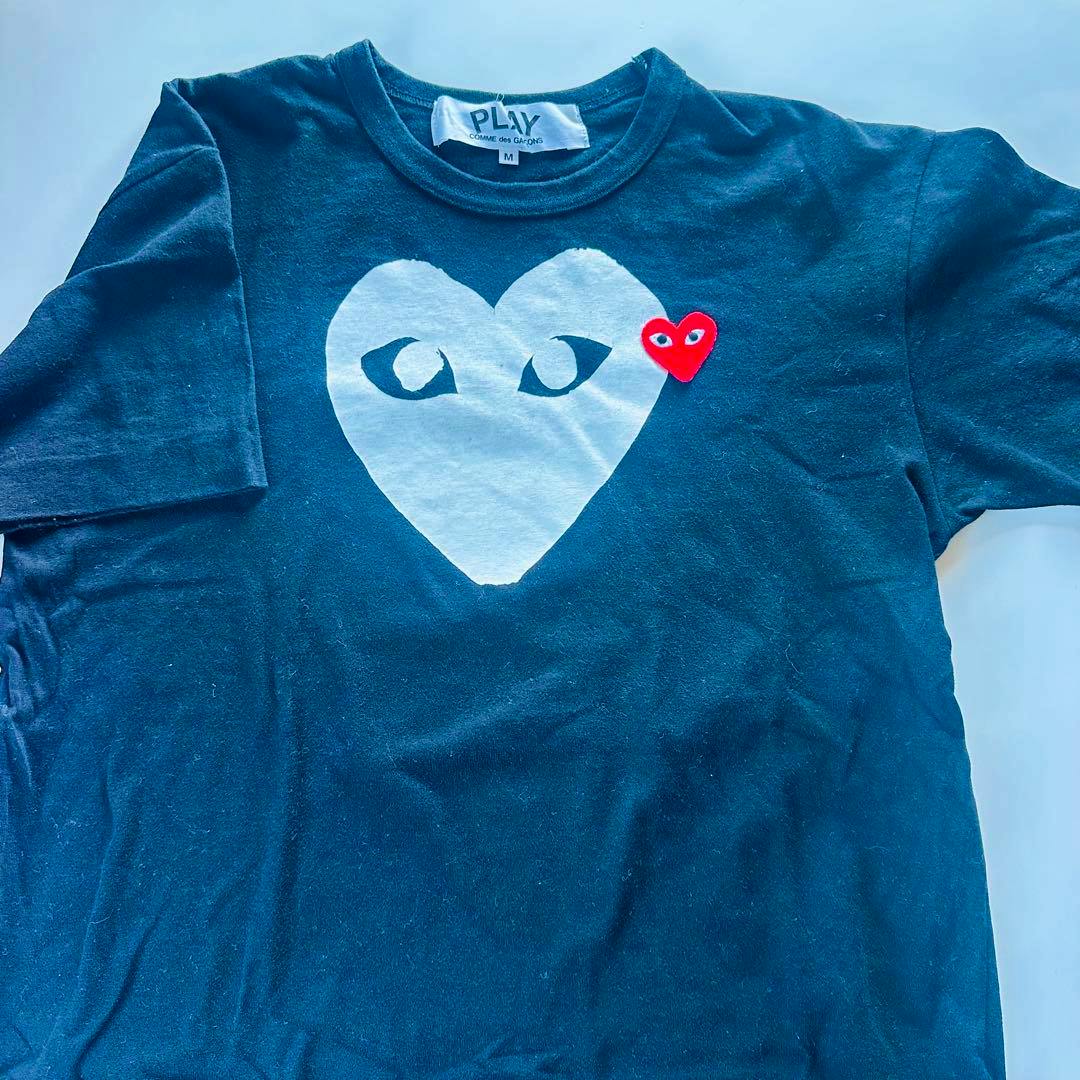 Black T‑shirt laid flat showing a large inverted white heart graphic and a small red heart patch.