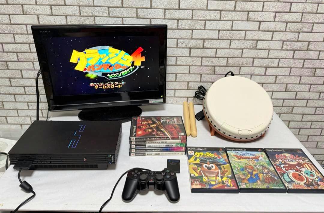 PlayStation 2 SCPH-39000 console on a table with a controller, memory card, multiple PS2 game cases, a drum controller, and a TV showing gameplay.