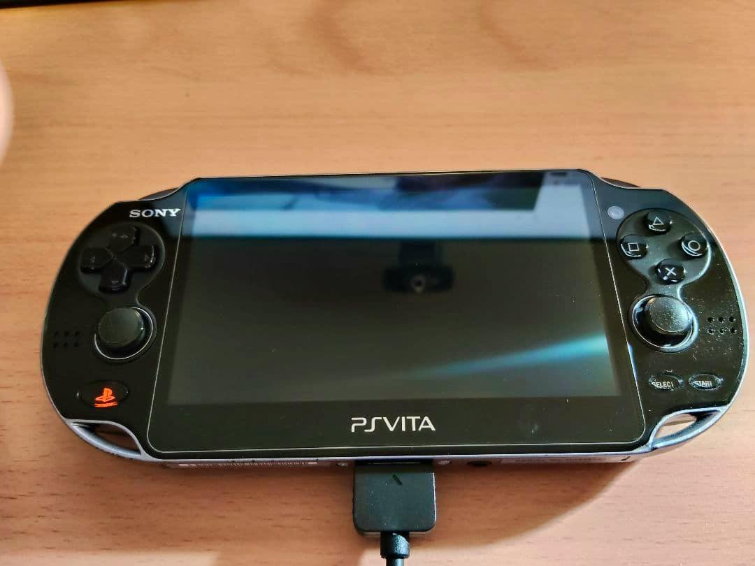 Front view of a black PS Vita PCH-1100 handheld console with screen, controls, and a charging cable connected.