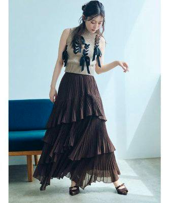 A model wearing a brown pleated tiered skirt shown full-length while standing indoors.
