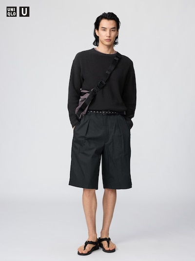 Male model standing facing camera wearing black wide pleated shorts with a black sweater and sandals.