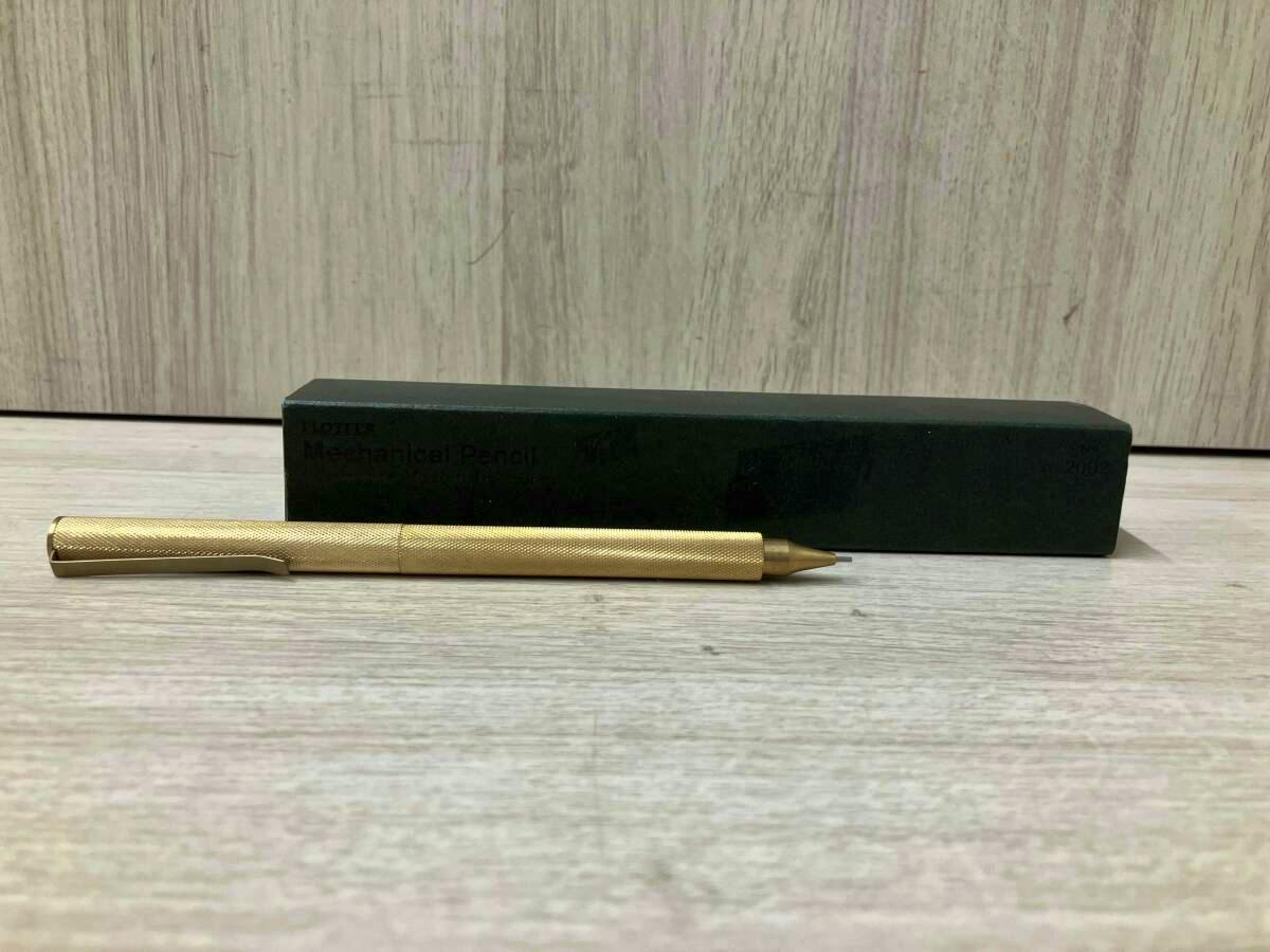 A gold PROTTER mechanical pencil shown lying in front of its dark rectangular box on a wood surface.