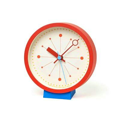 Front view of a round red table clock with a white face, stylized hands and hour markers, shown on a blue stand.