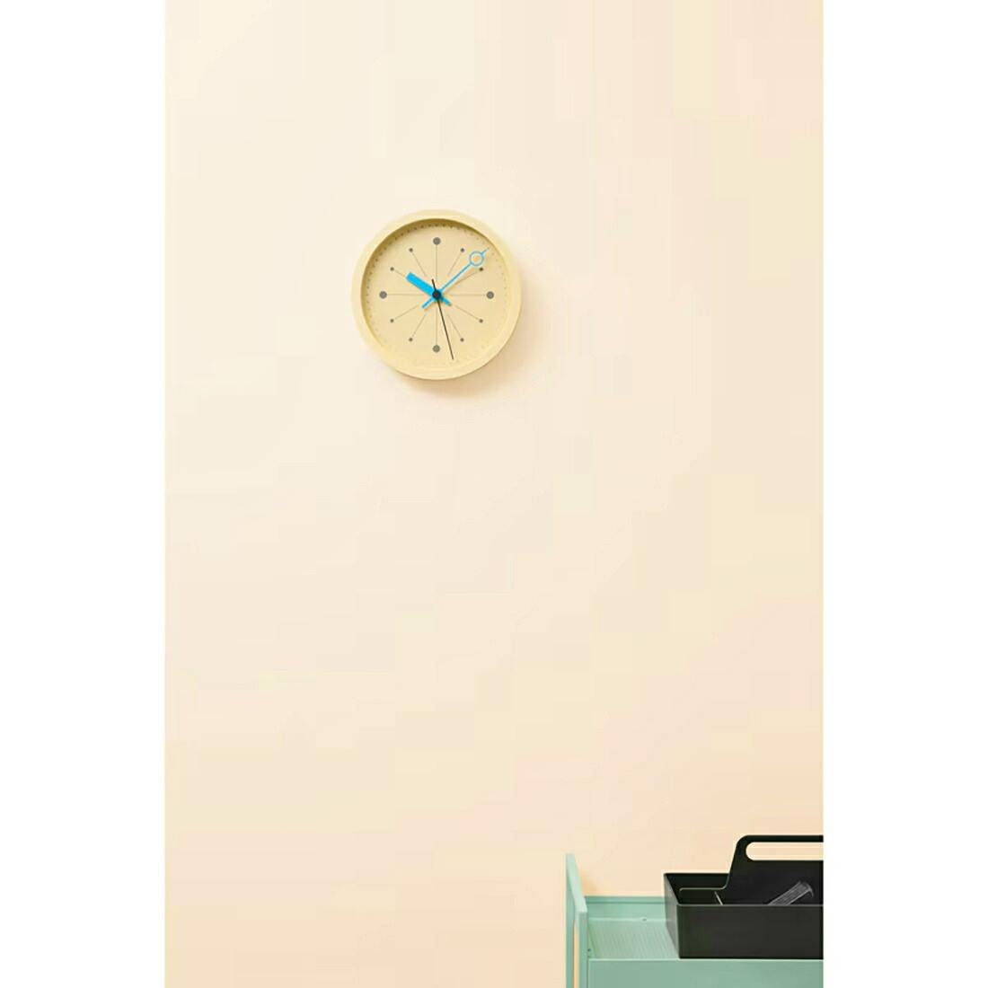 Round wall clock with blue hands mounted on a pale wall above a mint-colored shelf holding a black tray.