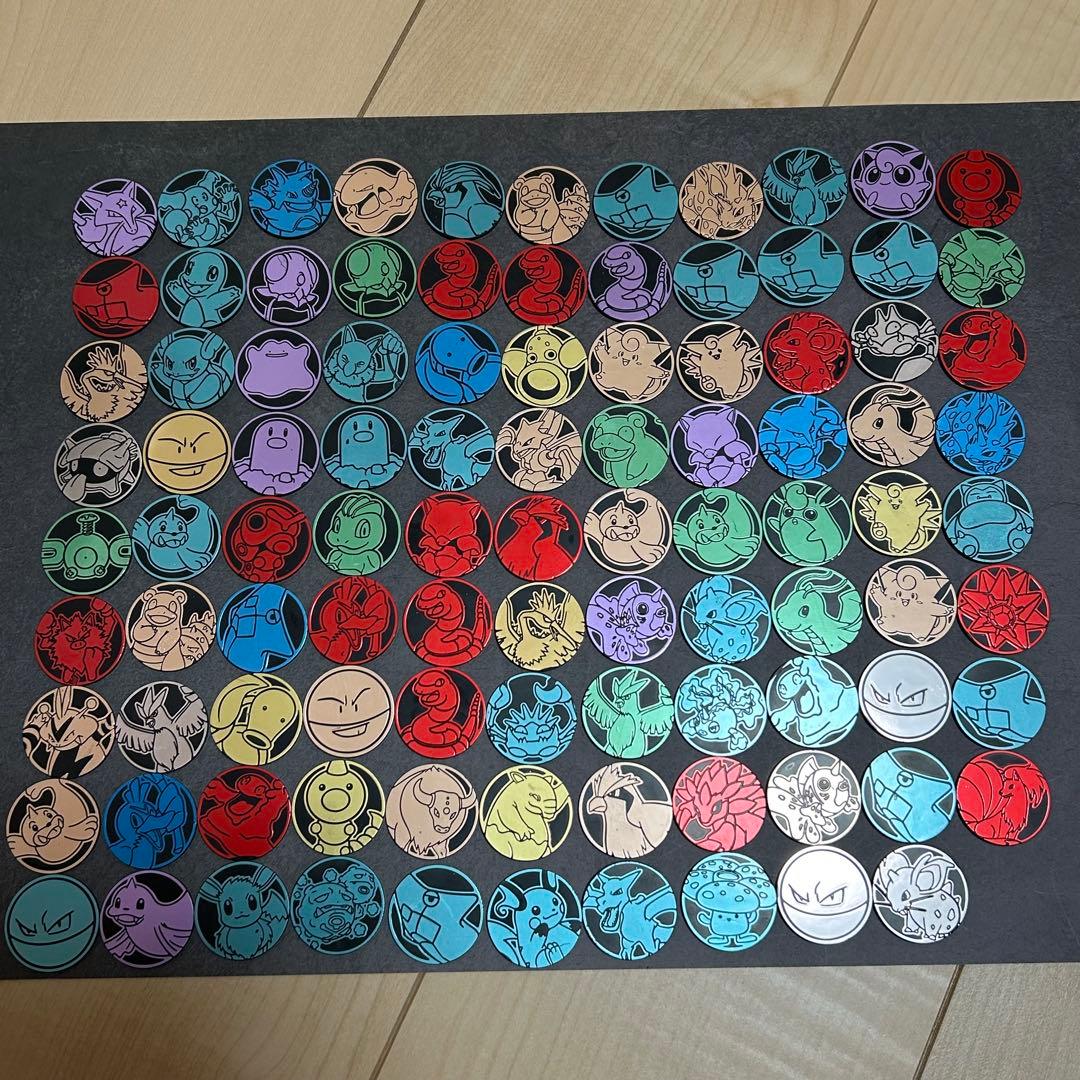 Overhead photo of many Pokémon-themed round metal coins laid out in rows on a dark surface.