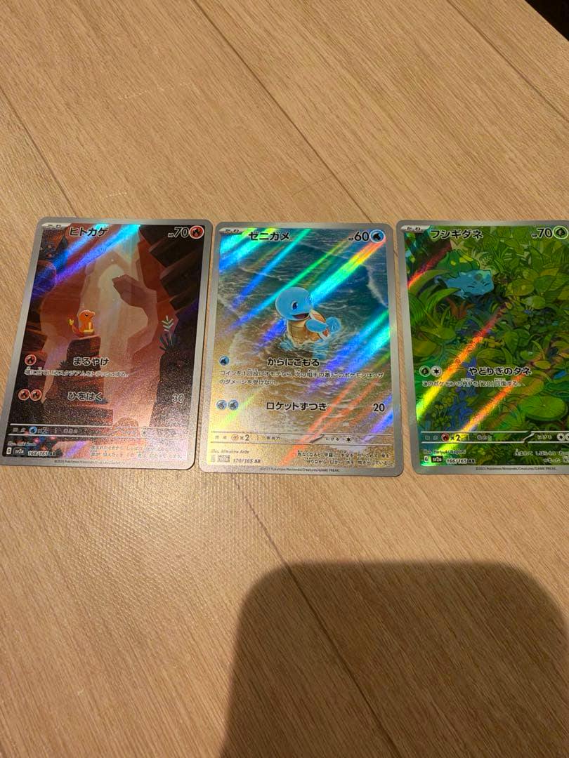 Three holographic Pokémon trading cards of the original starter trio laid side by side on a wooden surface.