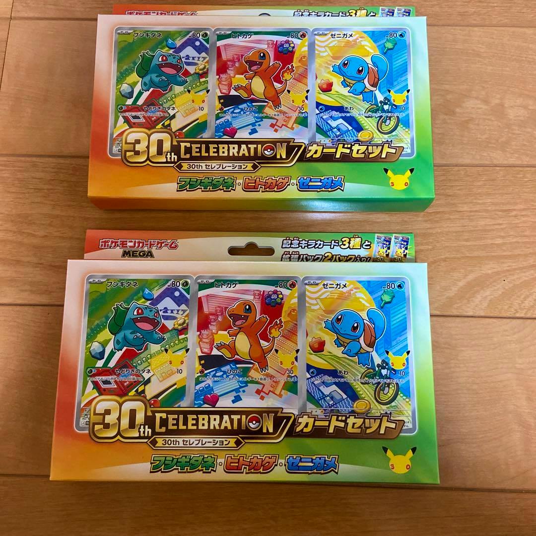 Two unopened Pokémon 30th Celebration card set boxes showing Bulbasaur, Charmander, and Squirtle on a wooden floor.