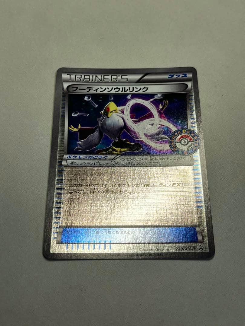 Photograph of the front of the フーディンソウルリンク Trainer Pokémon card with holographic artwork and a Pokémon Center campaign seal on a gray background.