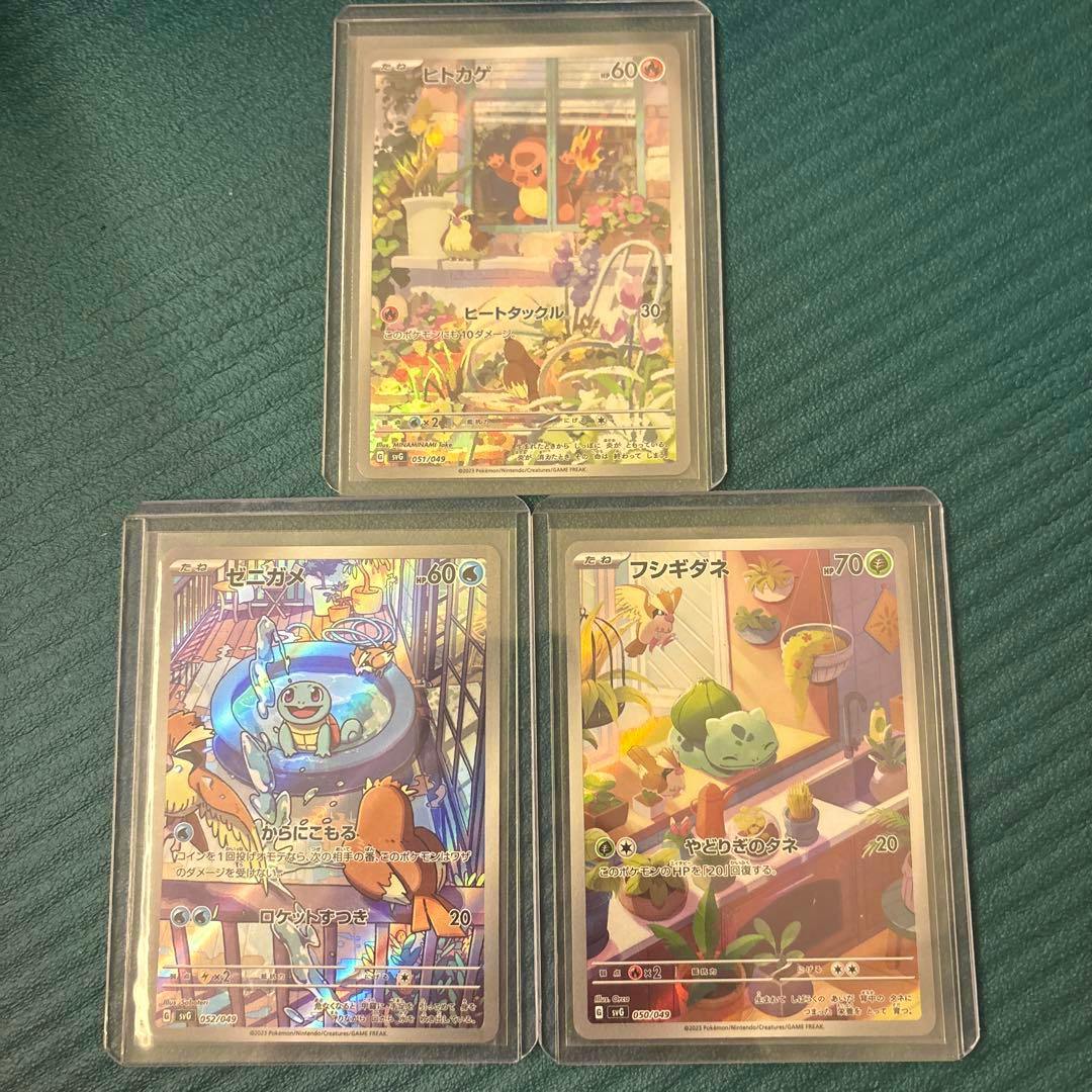 Three Pokémon trading cards from the listed set (ヒトカゲ, ゼニガメ, フシギダネ) shown in protective sleeves on a green surface.