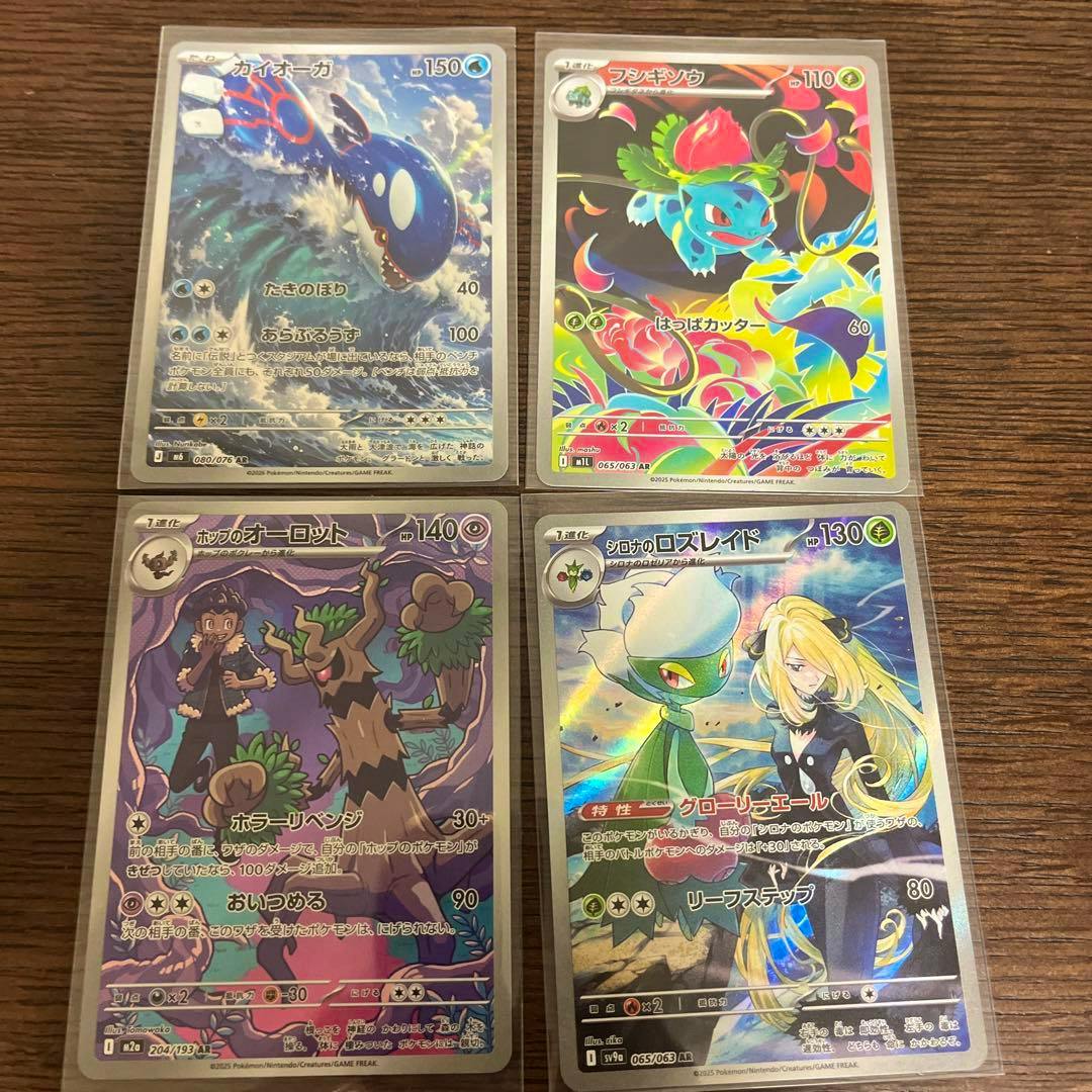 Four sleeved Pokémon trading cards showing their fronts arranged in a two-by-two grid on a wooden surface.