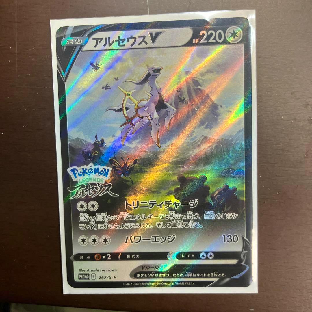 Holographic front of the Japanese “アルセウスV” Pokémon promo trading card shown in a clear sleeve on a dark wooden surface.