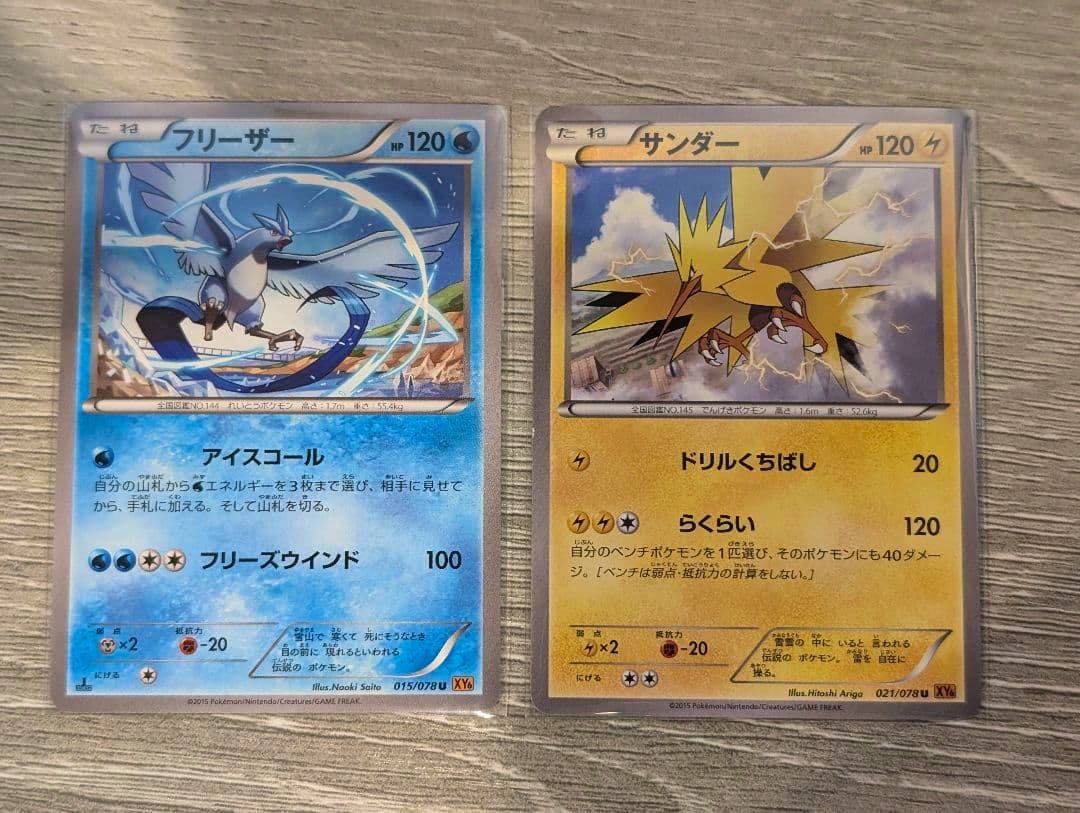 Fronts of two sleeved Japanese Pokémon trading cards laid side by side on a wooden surface, showing フリーザー on the left and サンダー on the right.