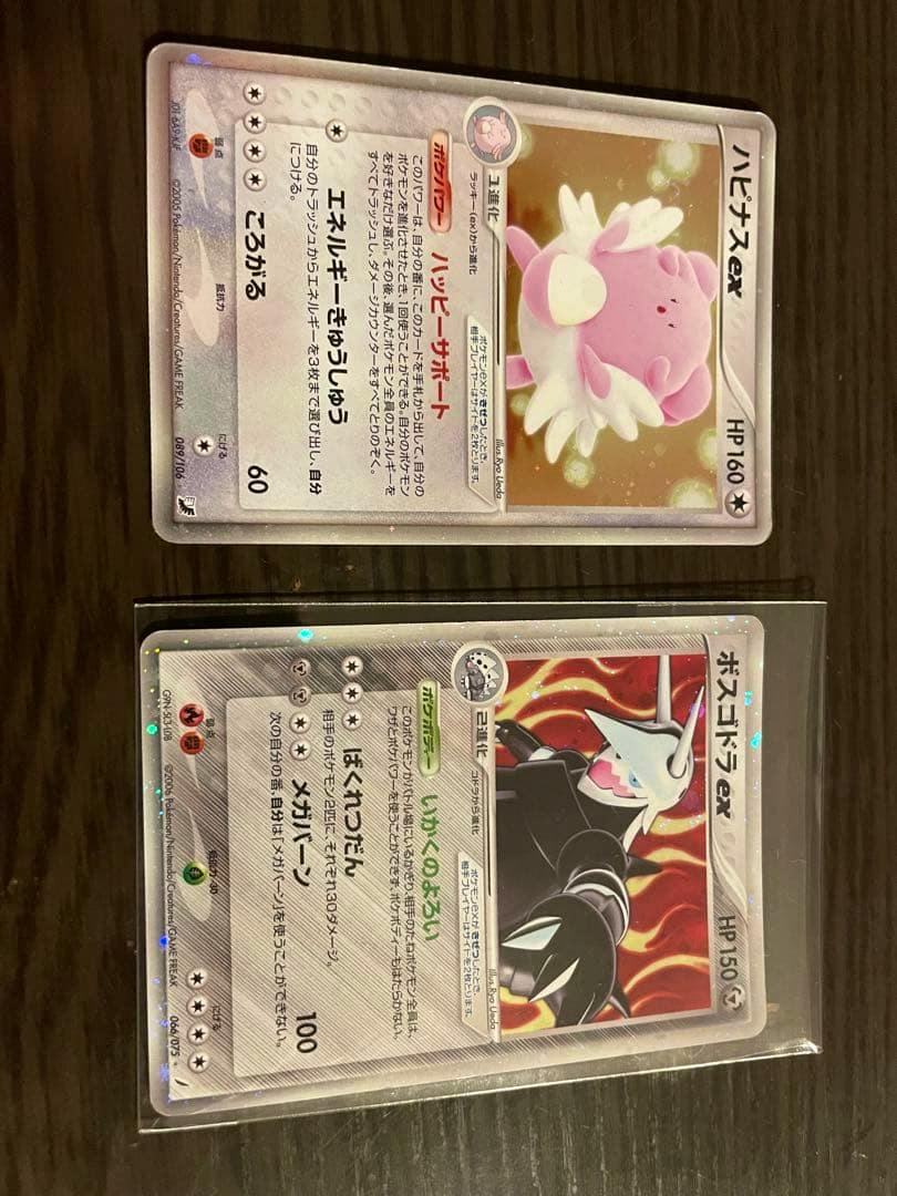 Two Pokémon trading cards (ハピナスex and ボスゴドラex) photographed face-up on a dark surface.