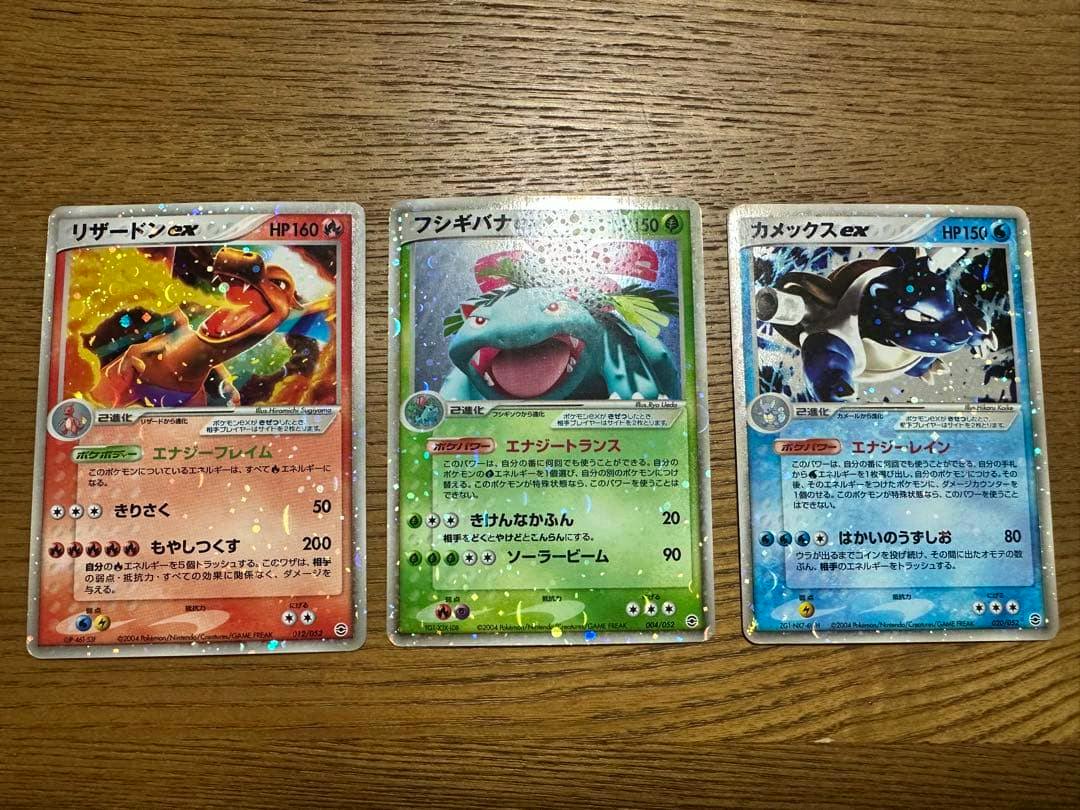 Fronts of three Pokémon trading cards — リザードンex, フシギバナ, and カメックスex — arranged side by side on a wooden surface.