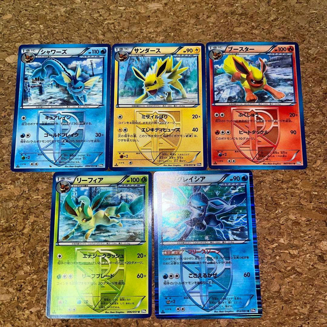 Five Pokémon trading cards showing Eevee evolutions (Vaporeon, Jolteon, Flareon, Leafeon, Glaceon) from the Plasma Team arranged on a cork surface.