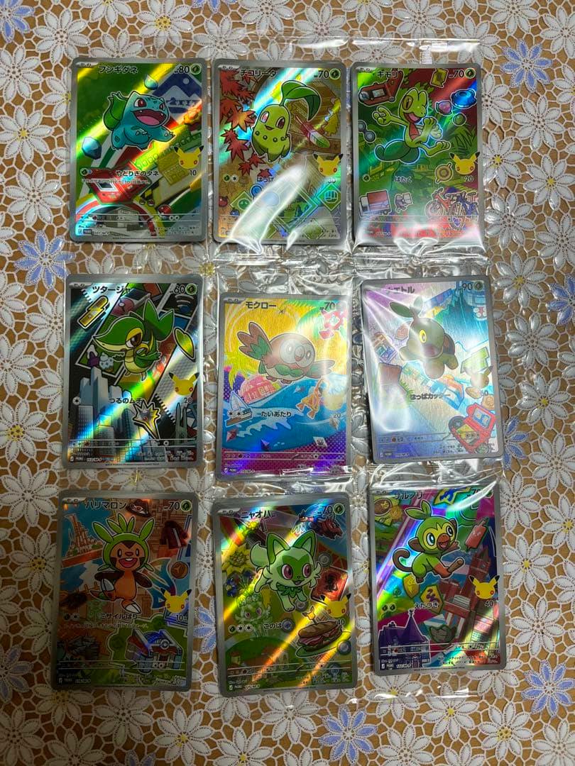 Nine Pokémon 25th‑anniversary promotional trading cards in individual sleeves arranged three-by-three on a patterned tablecloth.