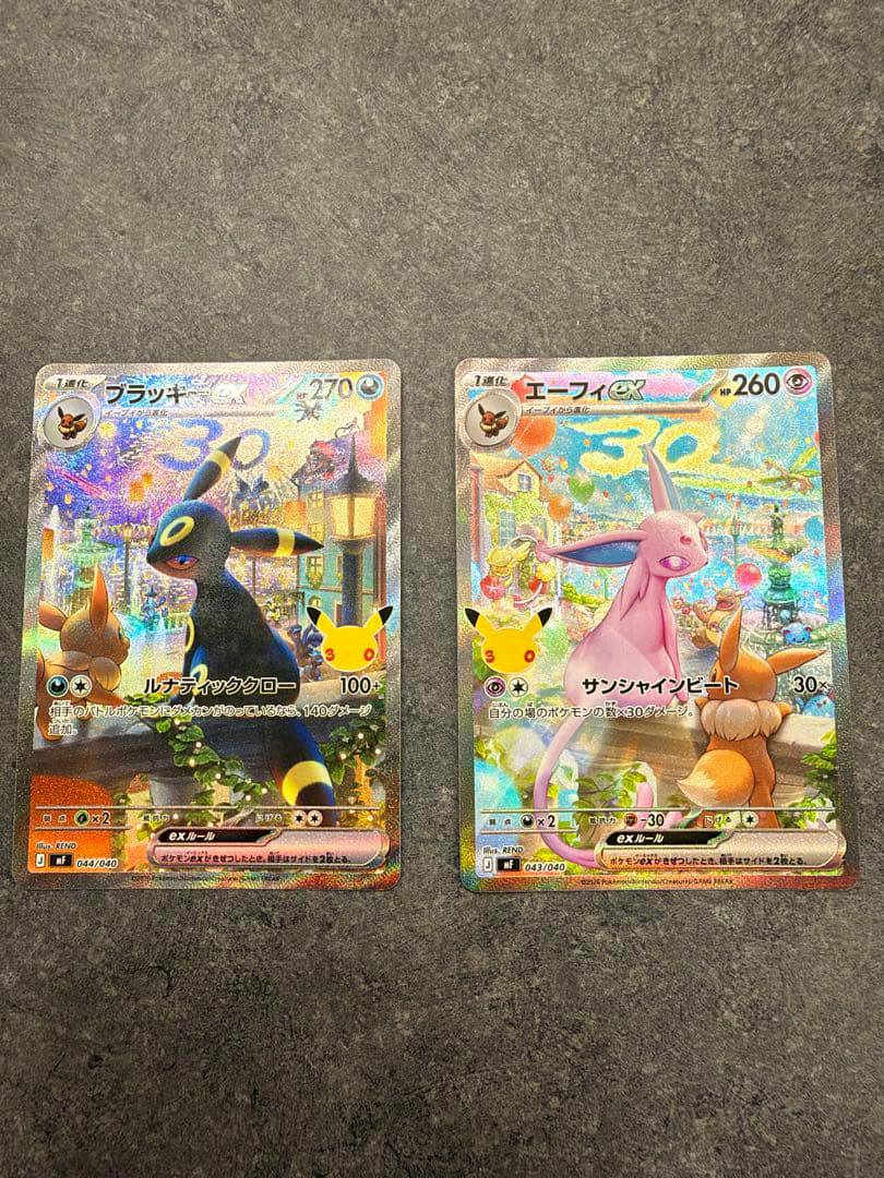 Photo of two Pokémon trading cards from the 30th anniversary premium deck set showing ブラッキーex (Umbreon ex) on the left and エーフィex (Espeon ex) on the right on a gray surface.