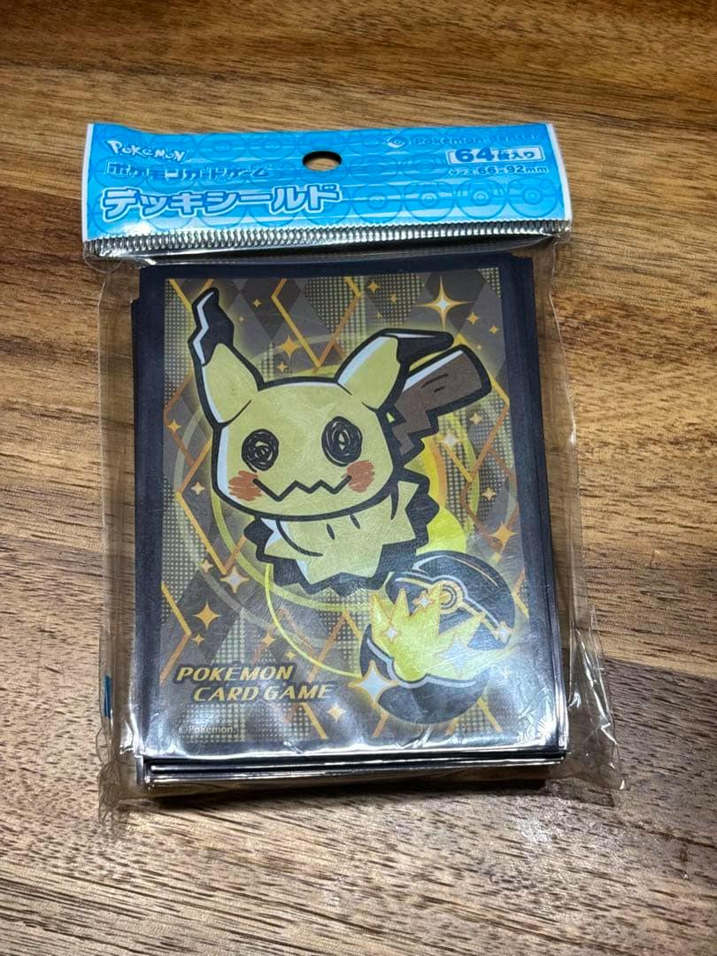 Packaged Pokémon card sleeves (Deck Shield) showing a Mimikyu design in clear plastic packaging on a wooden surface.