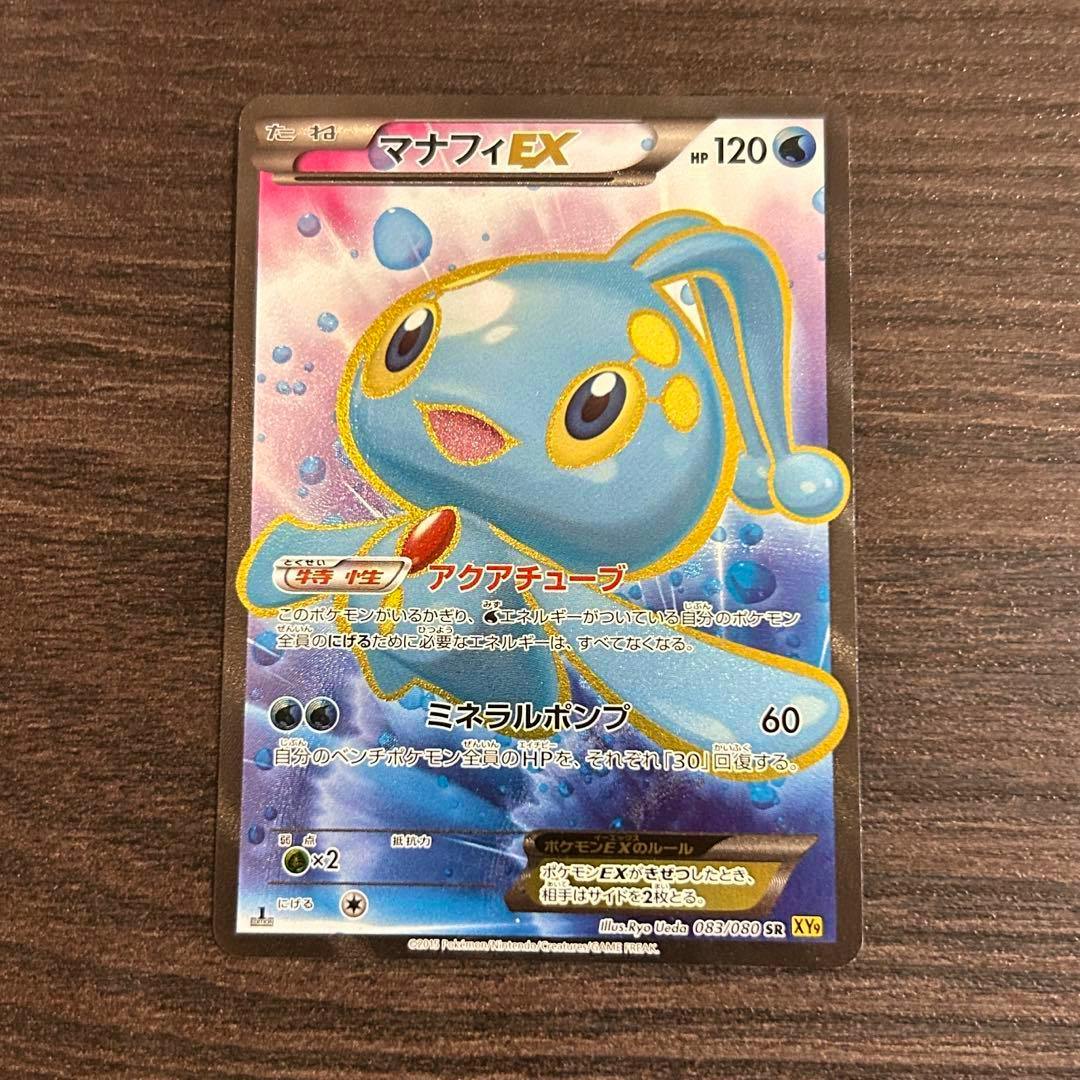 Front of a Japanese Manaphy EX Pokémon trading card (SR XY9 083/080) laying on a dark wood surface.