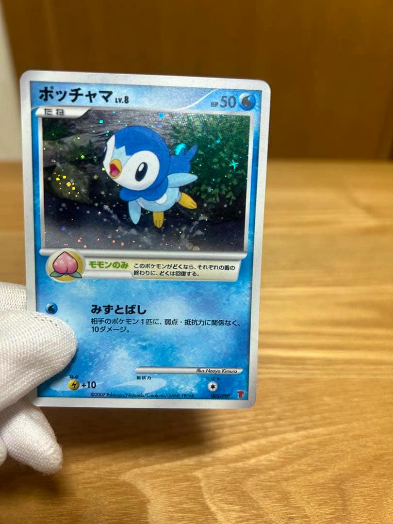 A Japanese Piplup (ポッチャマ) Pokémon trading card promo (003/PPP) held by a white-gloved hand over a wooden surface.