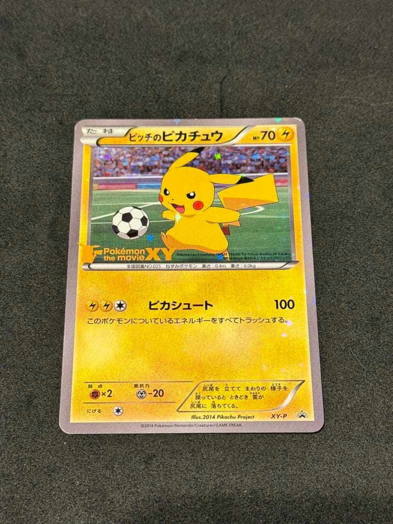 Front of the trading card “ピッチのピカチュウ” (promo XY-P) showing Pikachu with a soccer ball on a stadium background, placed on a black surface.