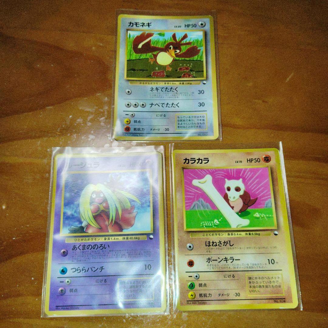 Three vintage Japanese Pokémon trading cards (カモネギ, ルージュラ, カラカラ) in clear sleeves laid on a wooden surface.