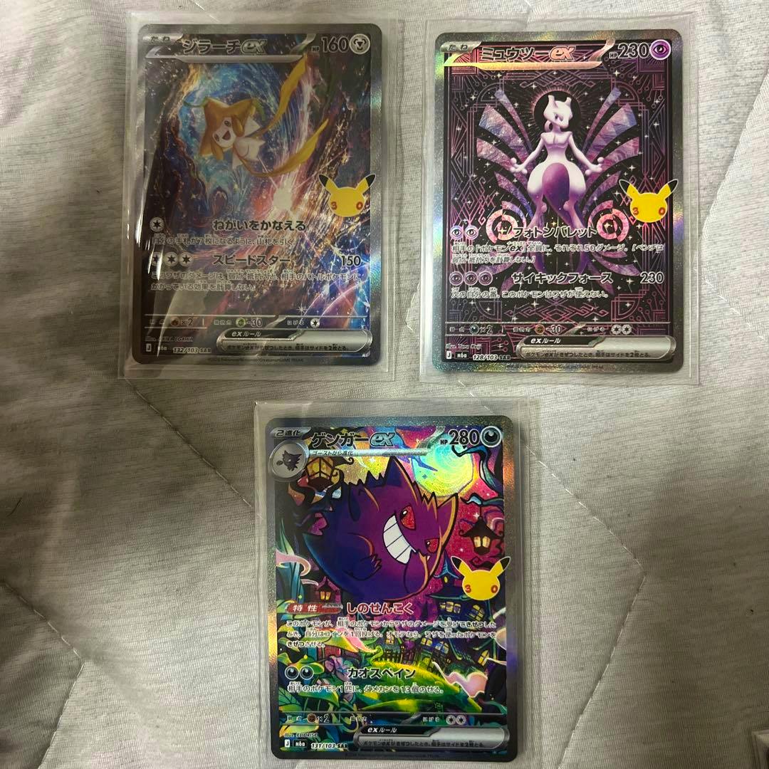 Three Pokémon trading cards (Jirachi, Mewtwo, and Gengar) in protective sleeves laid on a fabric surface.