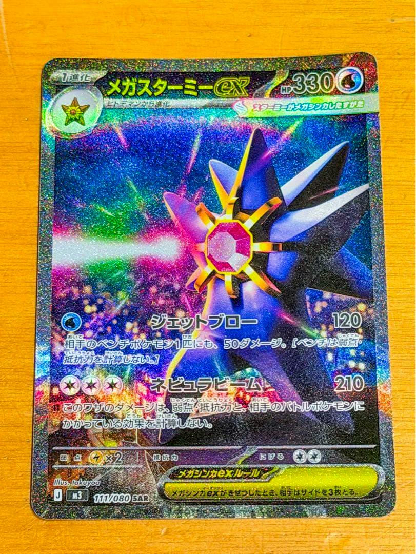 Photograph of the Pokémon trading card ポケモンカード メガスターミーex SAR 111/080 J M3 showing the holographic front of the card on a wooden surface.