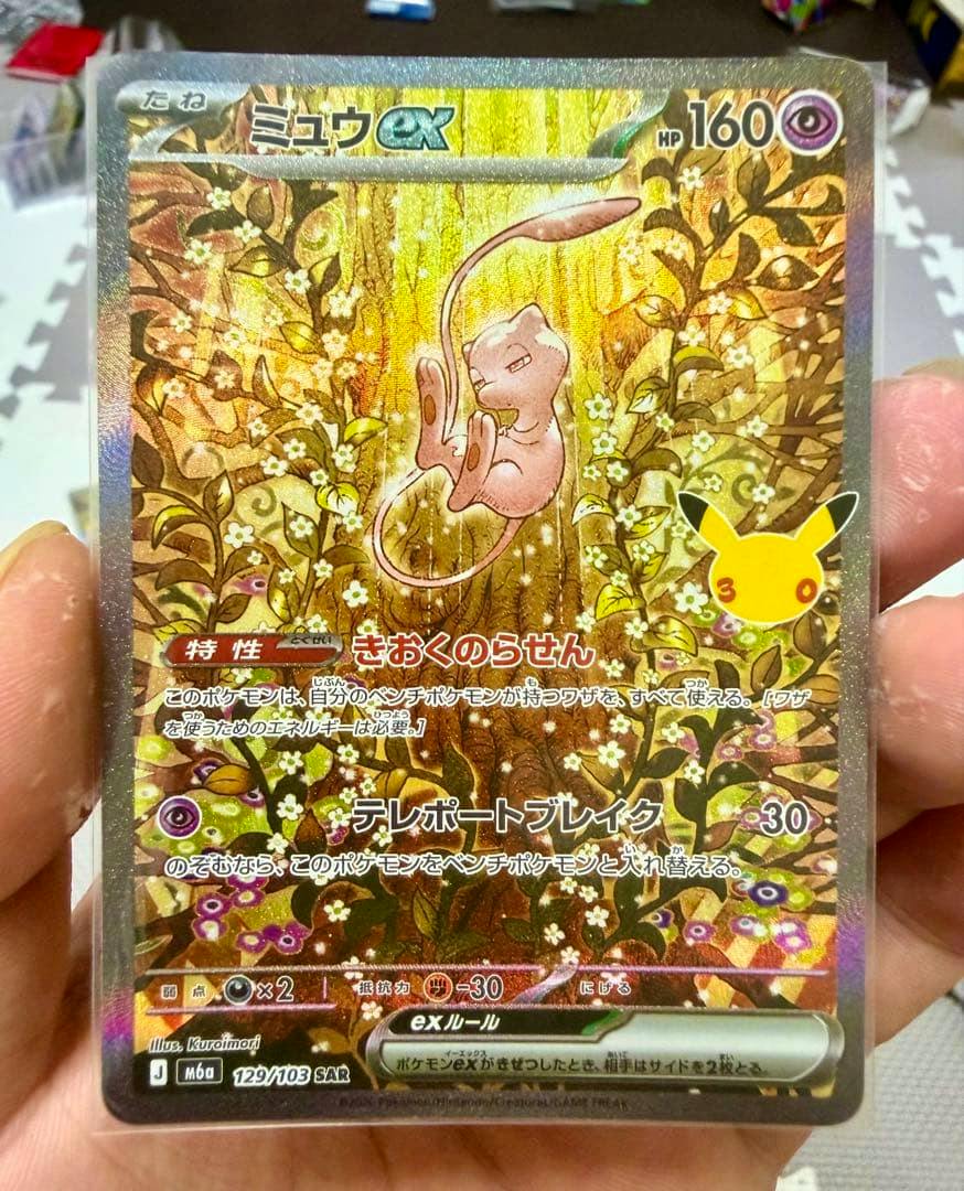 Photograph of the Pokémon trading card ポケモンカード ミュウex SAR 129/103 showing the full holo Mew ex artwork held in a hand.