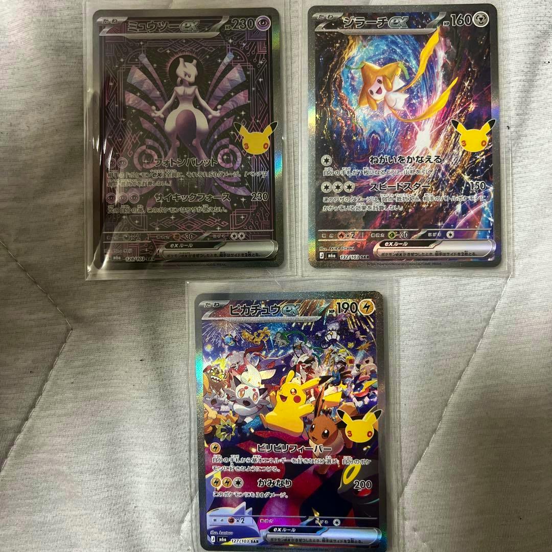 Three Pokémon trading cards (Mewtwo SAR, Jirachi SAR, and Pikachu SAR) in protective sleeves laid on a cloth surface.