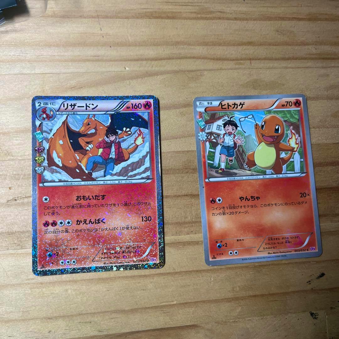 Two Pokémon trading cards showing Charizard (リザードン) and Charmander (ヒトカゲ) laid on a wooden surface.