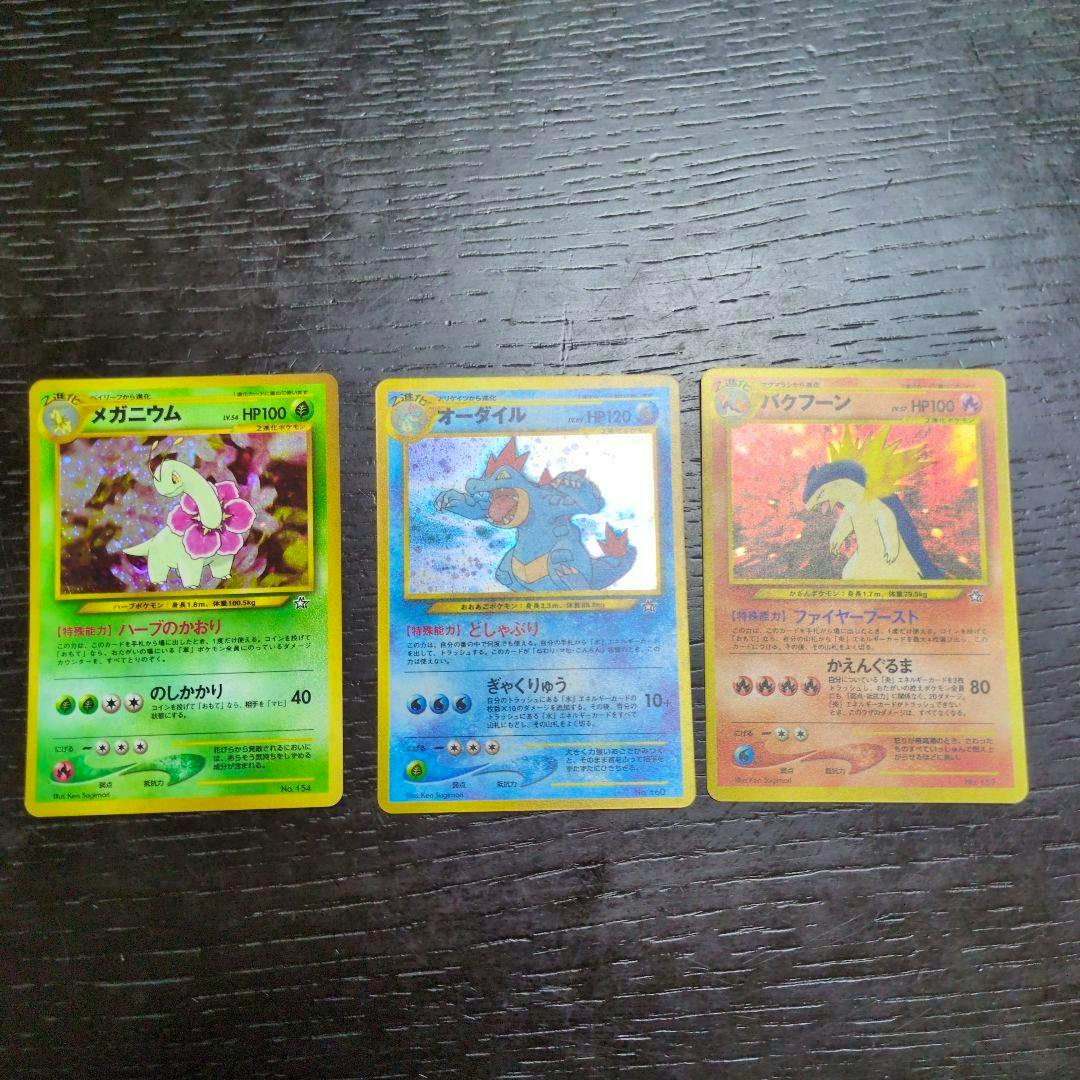 Three vintage Japanese Pokémon trading cards (メガニウム, オーダイル, バクフーン) laid side by side on a dark wood surface.