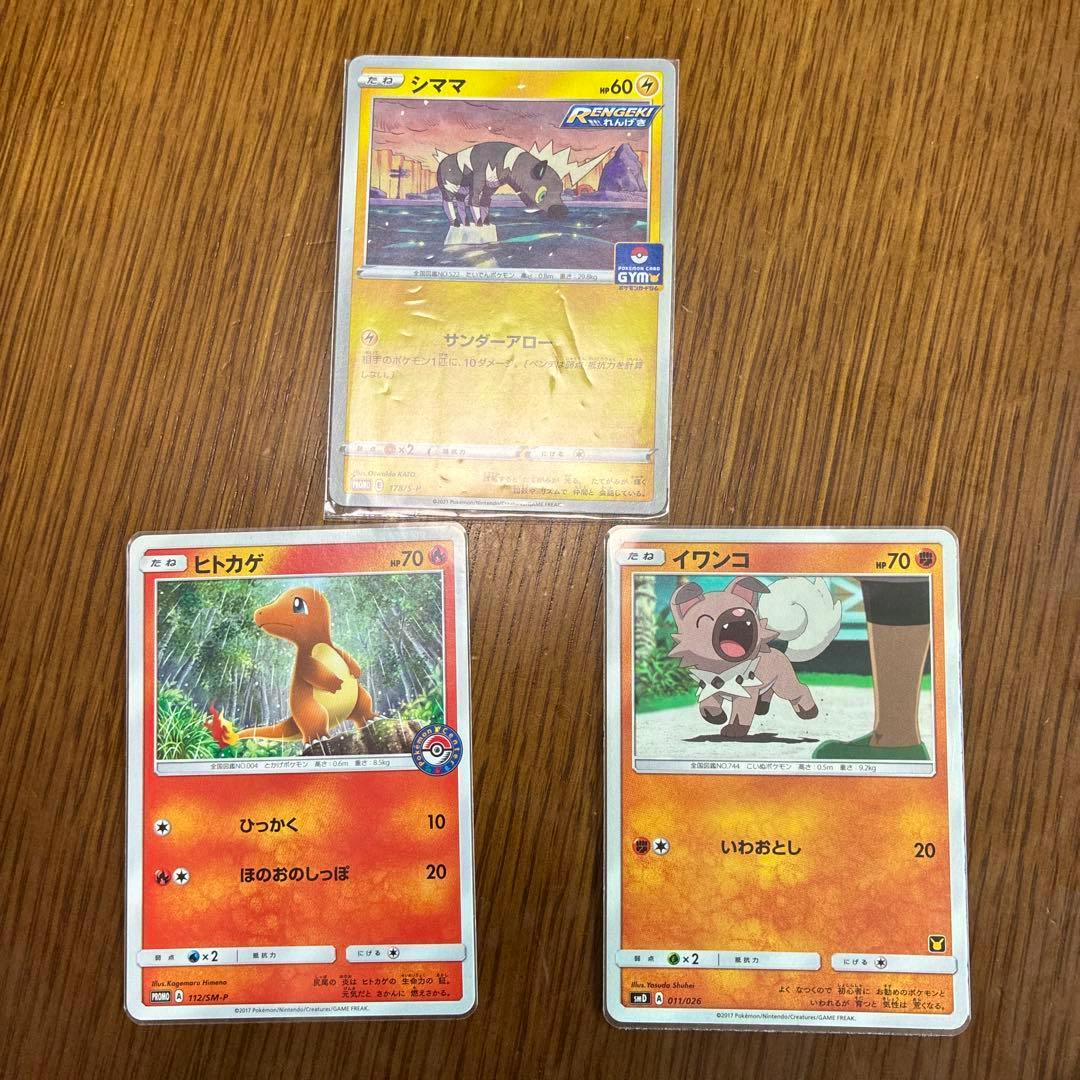 Three Pokémon promo trading cards (シママ, ヒトカゲ, イワンコ) laid on a wooden surface with the top card in a protective sleeve.