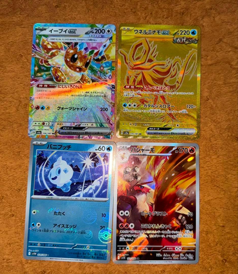 Four Pokémon trading cards laid out in a 2x2 arrangement on a wooden surface, showing a gold holo ウネルミナモ card among them.