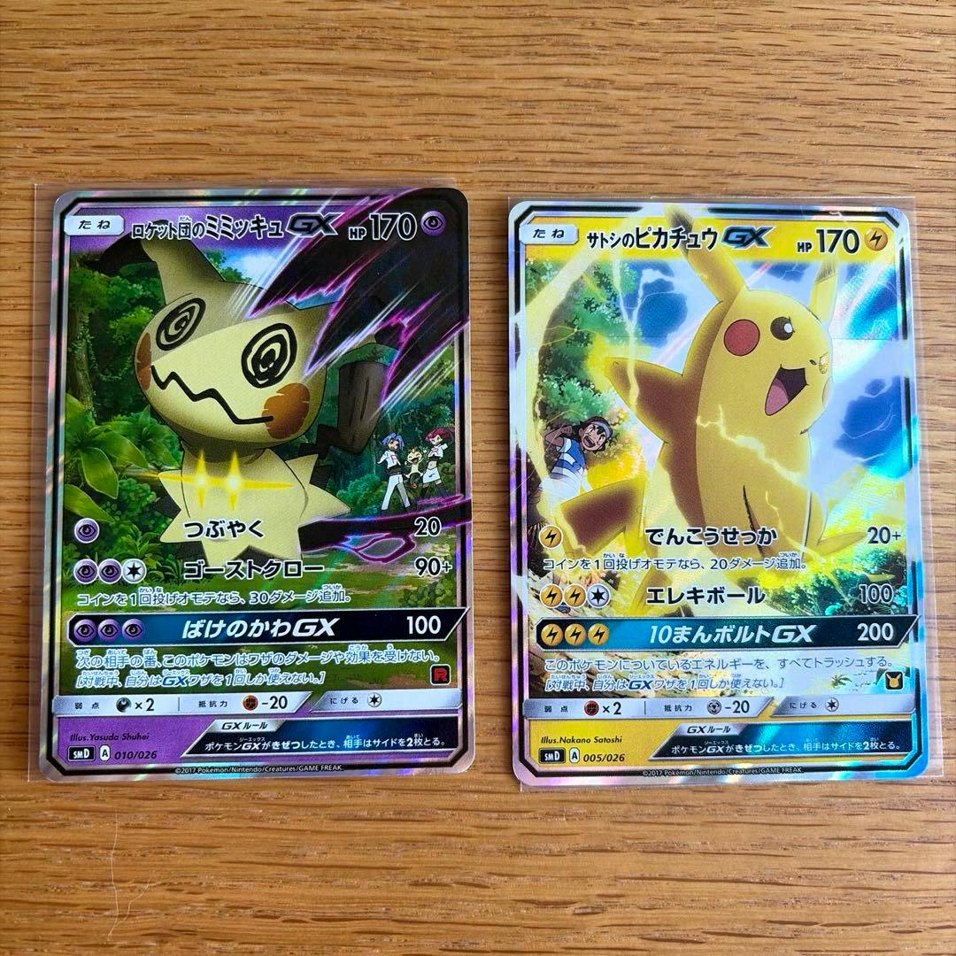 Front-facing photo of two Pokémon trading cards: Rocket Gang's Mimikyu GX (left) and Satoshi's Pikachu GX (right) on a wooden surface.