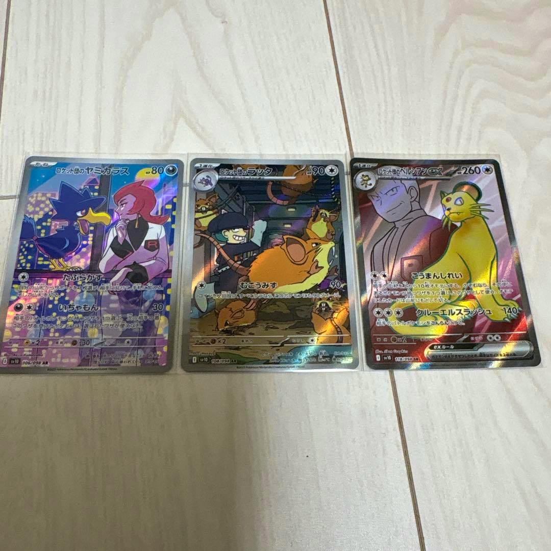 Three holographic Pokémon trading cards in sleeves laid on a wood floor showing Team Rocket Honchkrow, Raticate, and Persian ex.