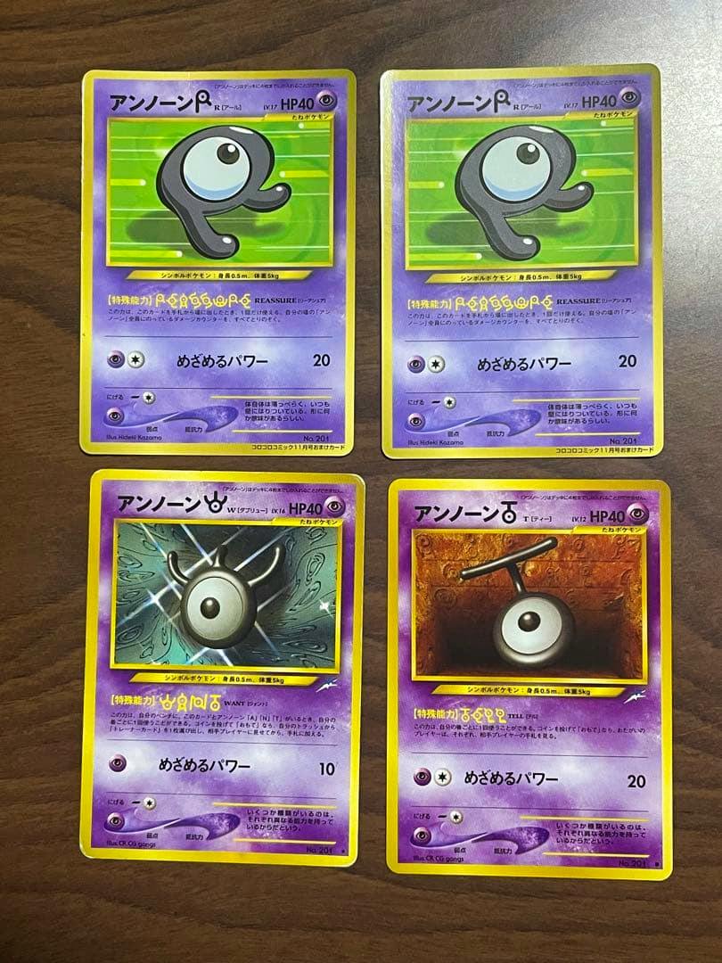 Four Unown Pokémon trading cards arranged in a 2x2 grid on a wooden surface, showing the front artwork and text.