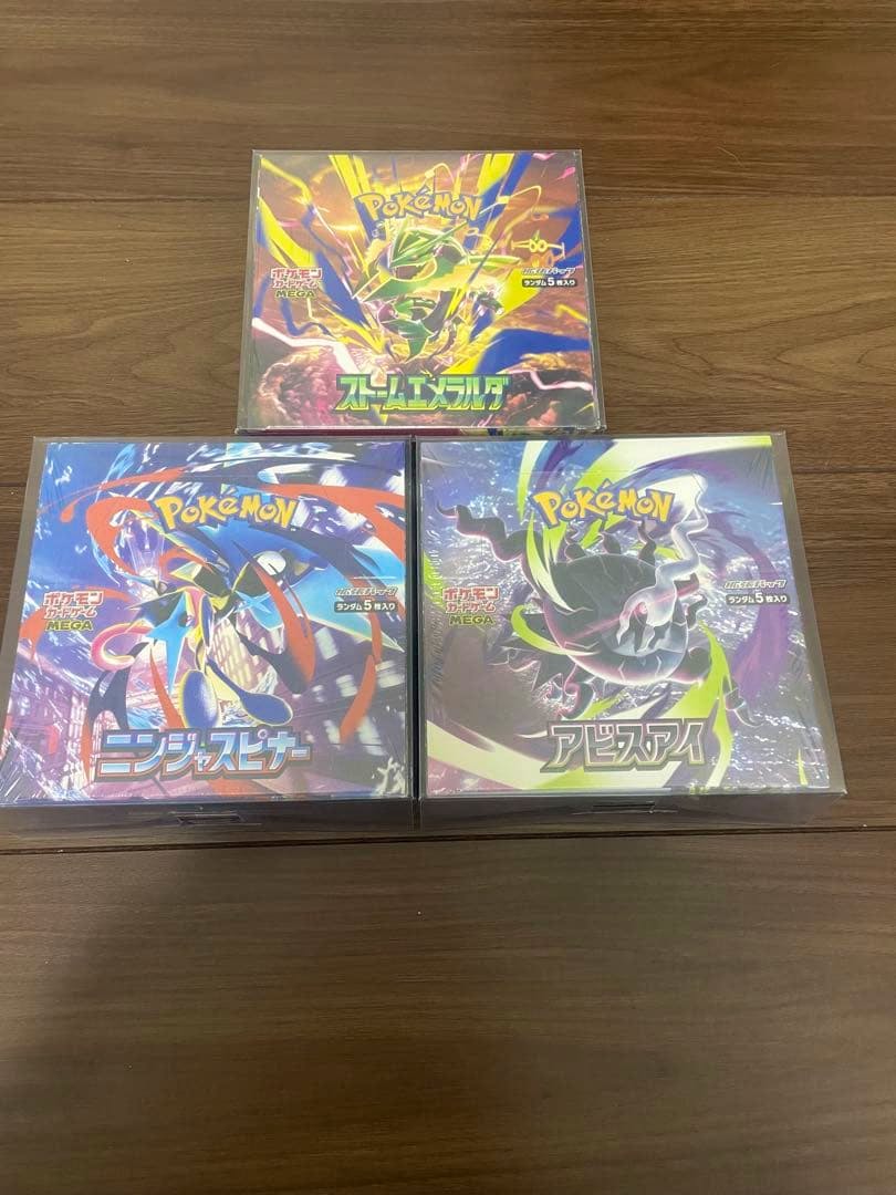 Three shrink-wrapped Pokémon card boxes—Storm Emerald (top), Ninja Spinner (bottom left), and Abyss Eye (bottom right)—displayed on a wooden surface.