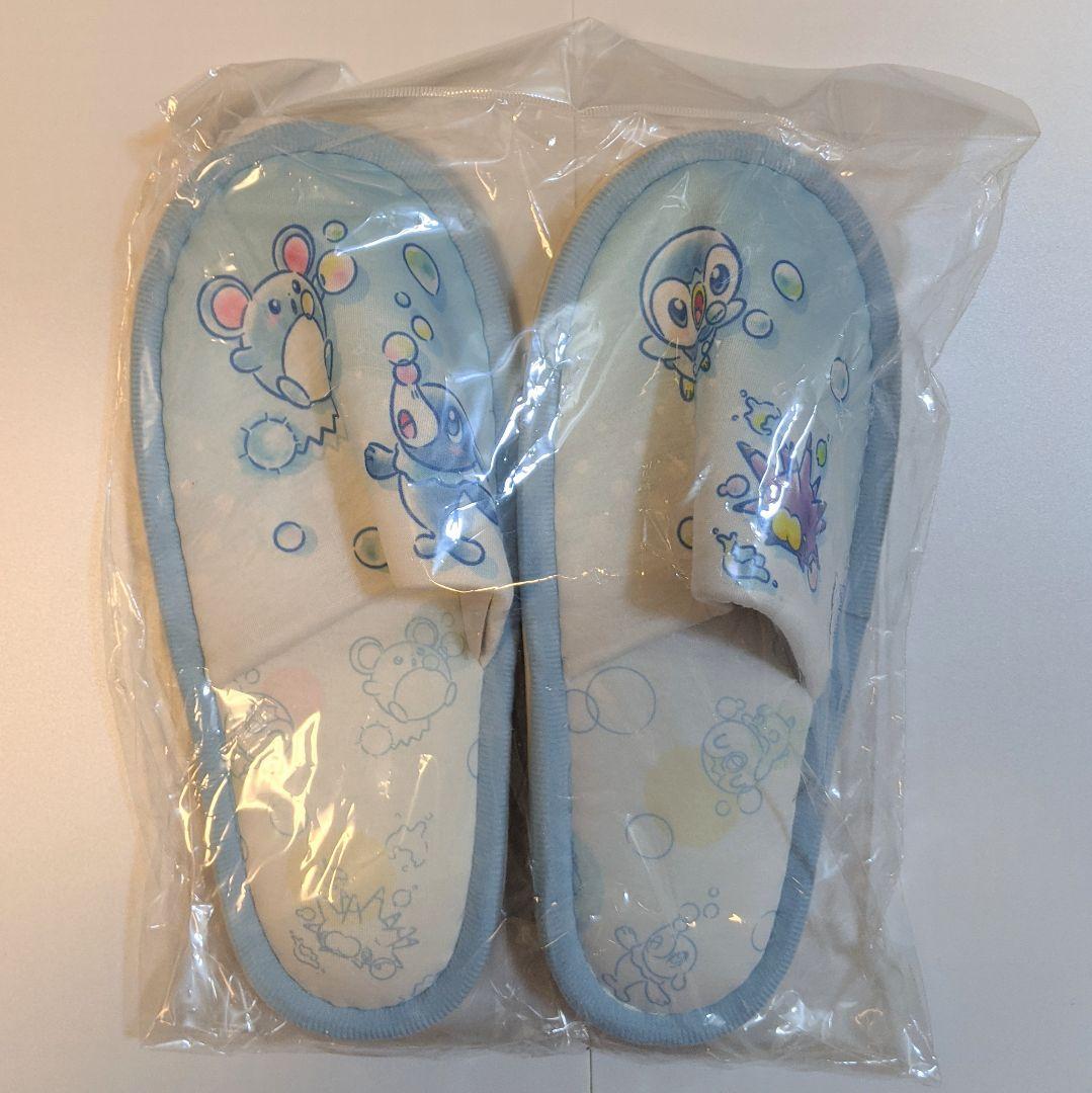 Top-down photo of a pair of light-blue Pokémon Center "bubbly hour" slippers with Piplup and bubble-themed illustrations inside plastic packaging.