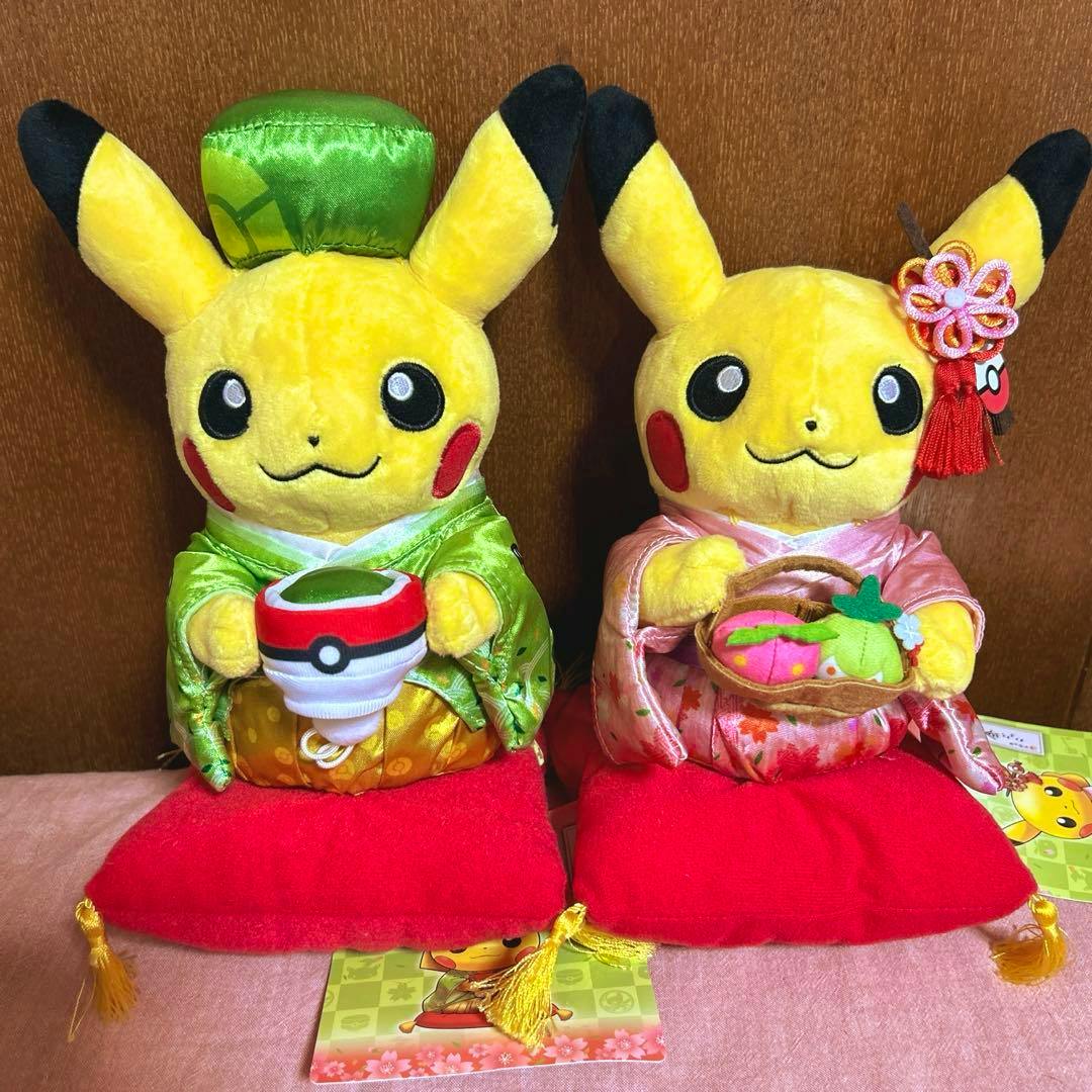 Two Pikachu plush toys in green and pink kimono seated on red cushions, each holding tea-themed props.