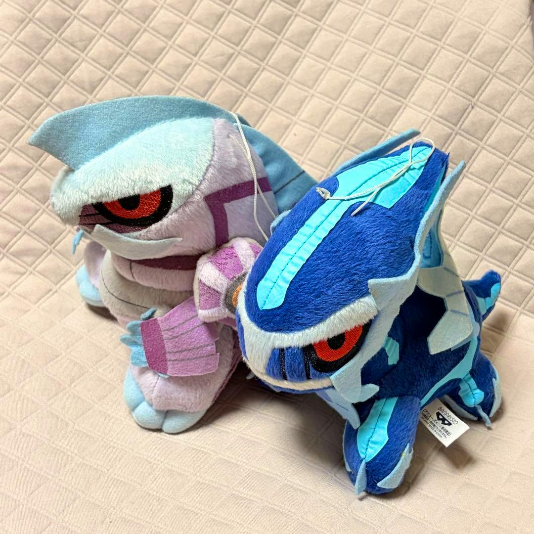 Photo of two Pokémon plush toys, Dialga (blue) and Palkia (pink), placed on a quilted beige surface.