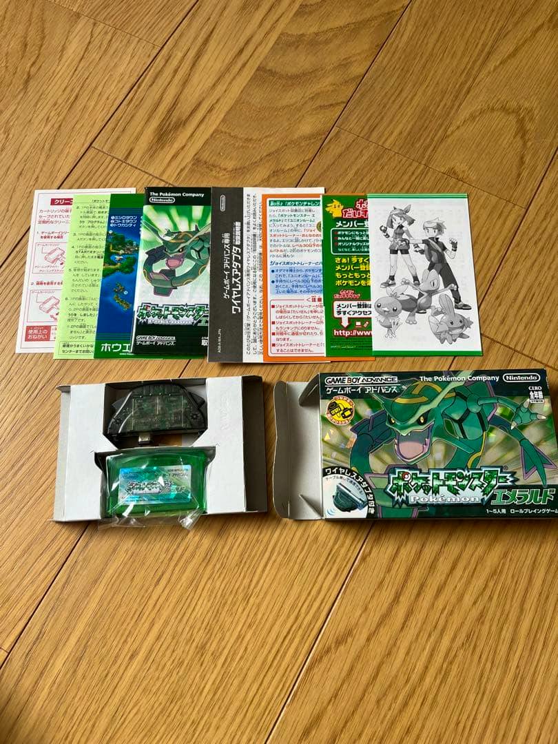 Box, green Game Boy Advance cartridge, wireless adapter, and paper manuals for Pocket Monsters Emerald laid out on a wooden floor.